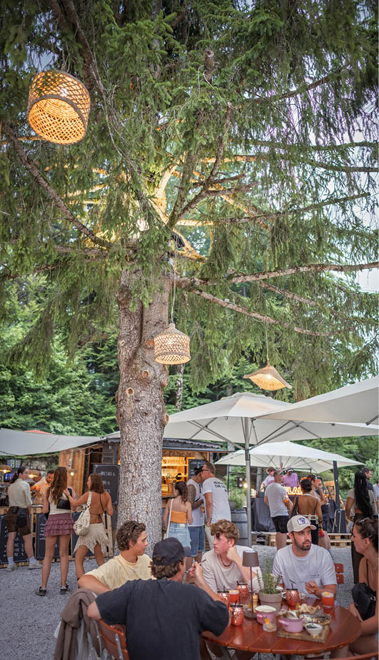 Gastgarten vom Almstadl am Schwarzensee. In den Bäumen hängen Bastlampen, darunter weiße Sonnenschirme und viele Menschen.