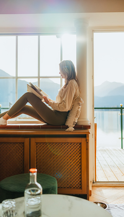 Eine Frau liest ein Buch auf der Fensterbank. Dahinter die Wolfgangseekulisse