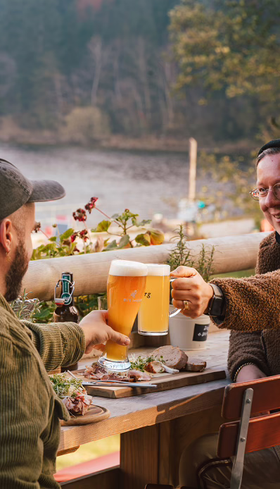 Zwei Männer stoßen auf einer Seeterrasse mit Bier an.